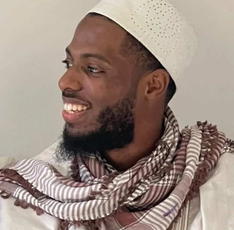 Hamza Abdul-Tawwaab, smiling and looking off to one side, wearing a white kufi and a brown-and-white patterned keffiyeh, against a plain pale wall.